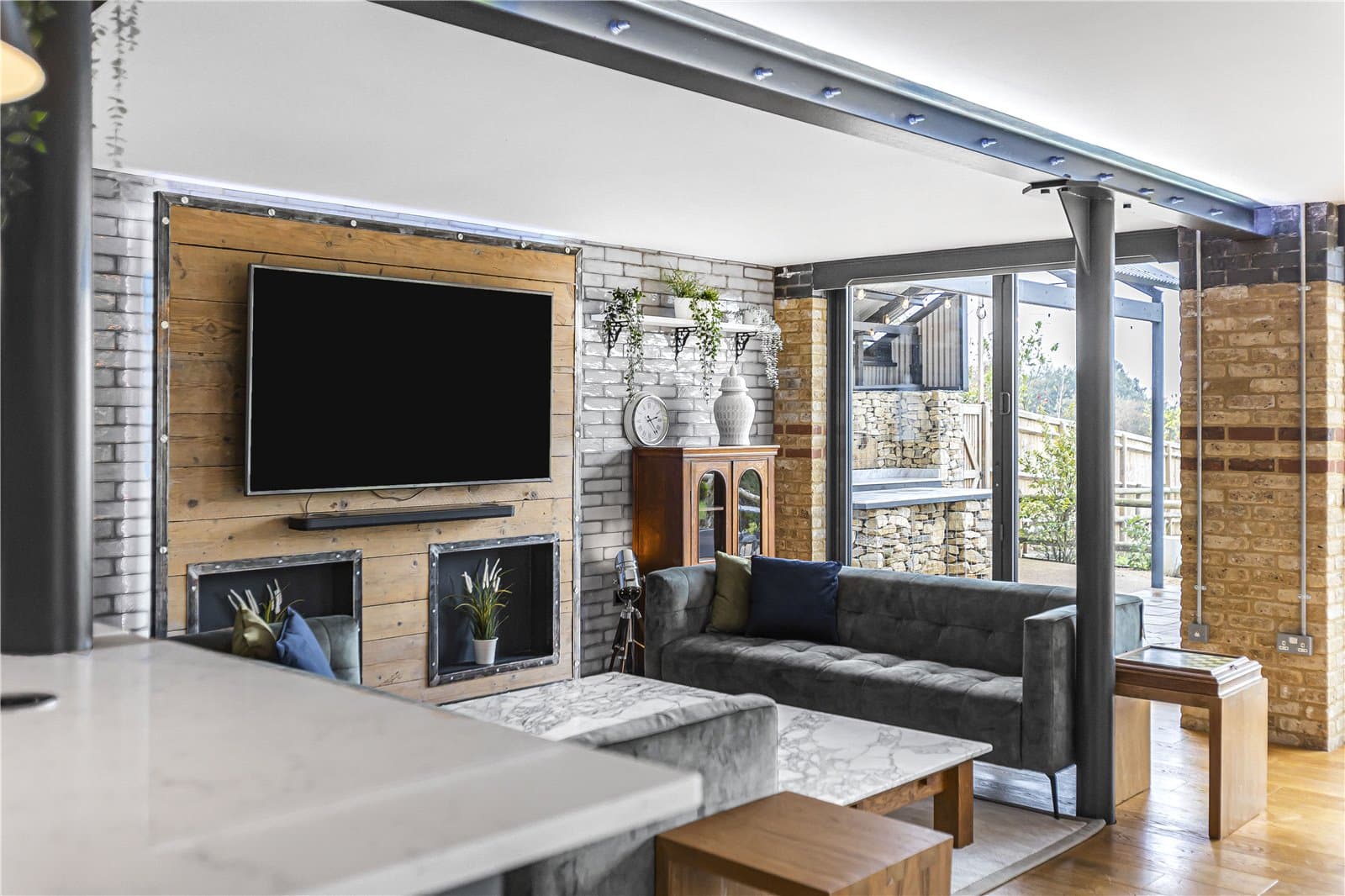 Open plan living area with television wall, exposed brick detailing and full height glazing overlooking the garden