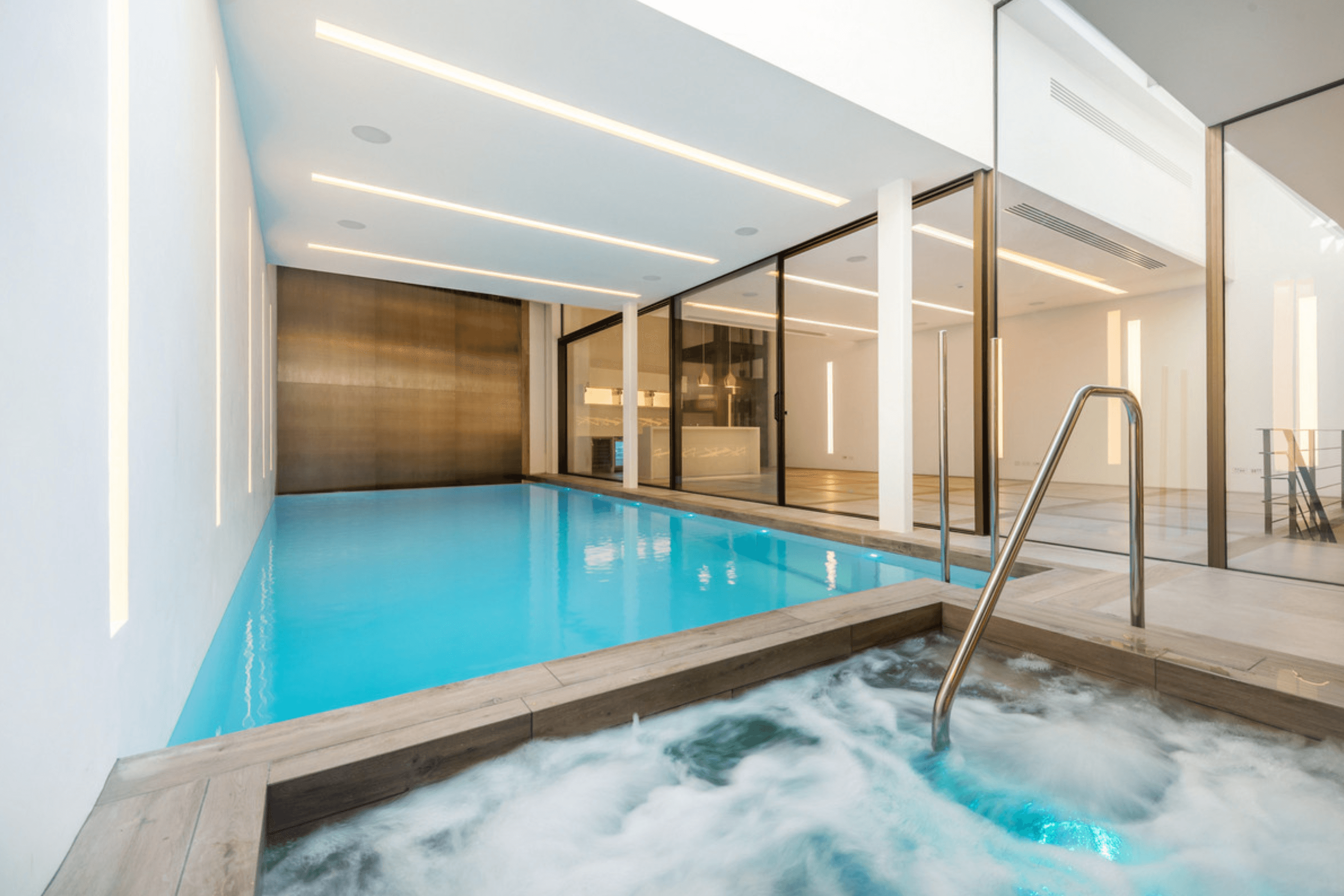 Indoor infinity pool with integrated jacuzzi set beneath recessed ceiling lighting and floor to ceiling glazing.