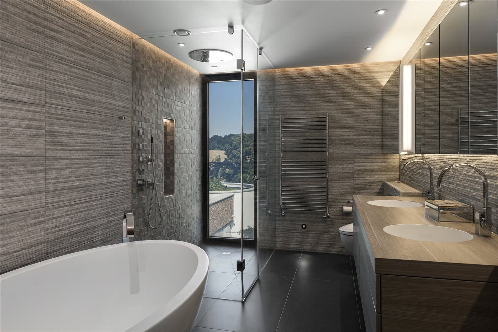 A bathroom with a freestanding oval bath, textured stone effect wall tiling, a walk in shower with an overhead rain shower head, and a double vanity unit with timber countertop. A narrow window offers a view over the surrounding countryside. The Warren, Radlett, Hertfordshire, listed with Prime Property.