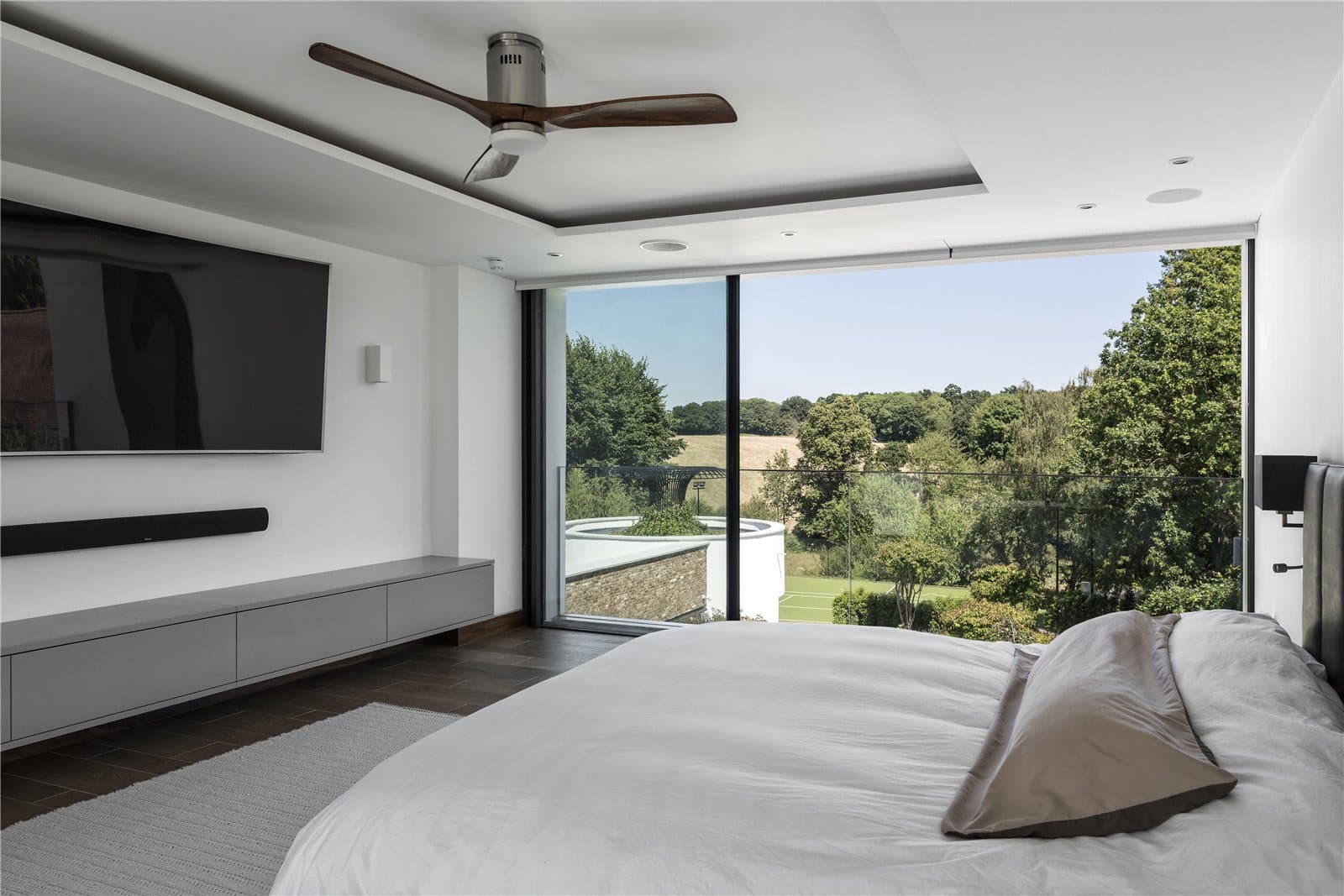 A bedroom with a wall mounted television, dark wood flooring and a ceiling fan, with full height sliding glass doors opening onto a balcony overlooking the tennis court and surrounding countryside. The Warren, Radlett, Hertfordshire, listed with Prime Property.