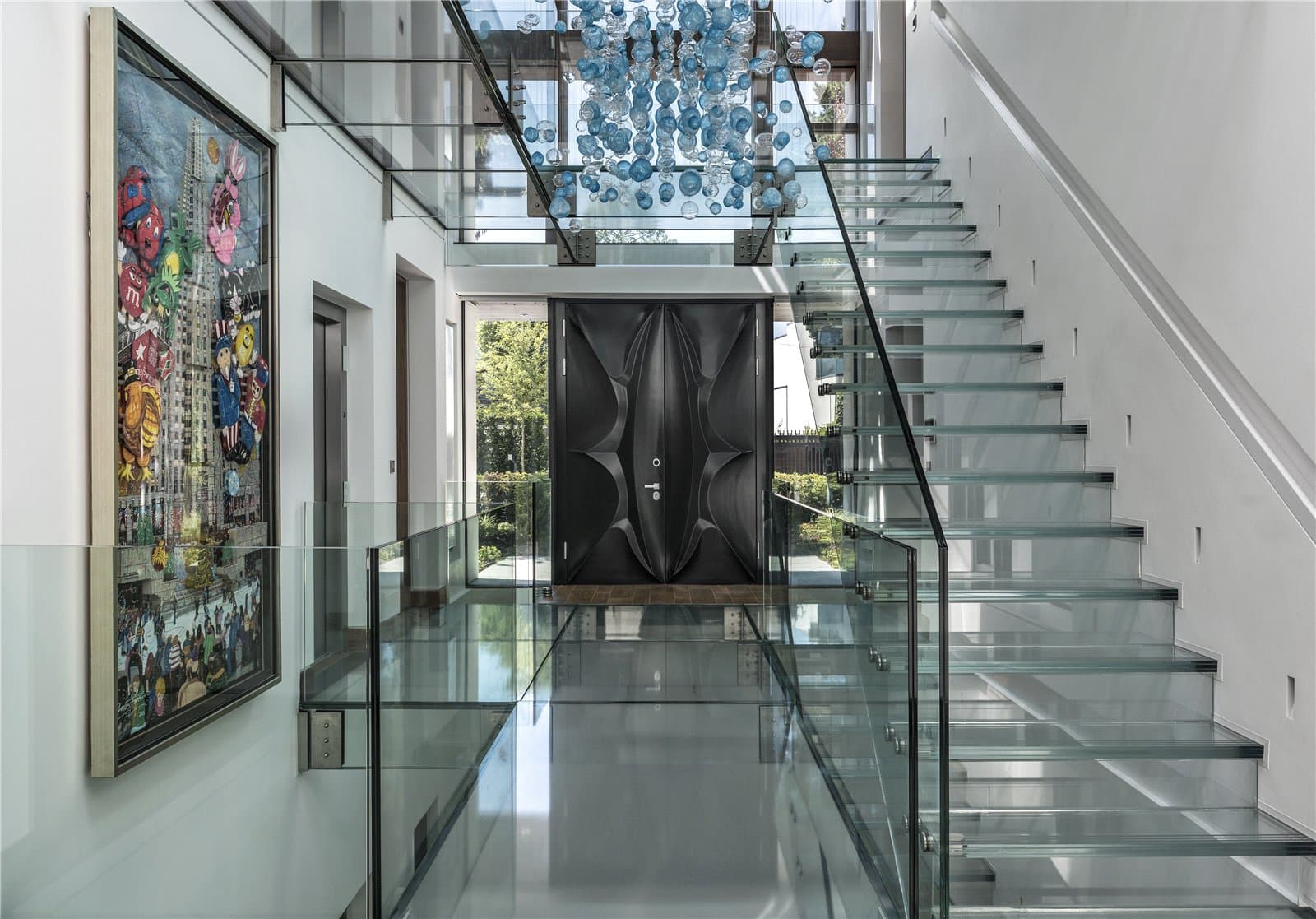 A glass staircase with a glass balustrade rises alongside a double height entrance hall, with a sculpted dark front door visible at ground level and a hanging glass bead light fixture above. The Warren, Radlett, Hertfordshire, listed with Prime Property.