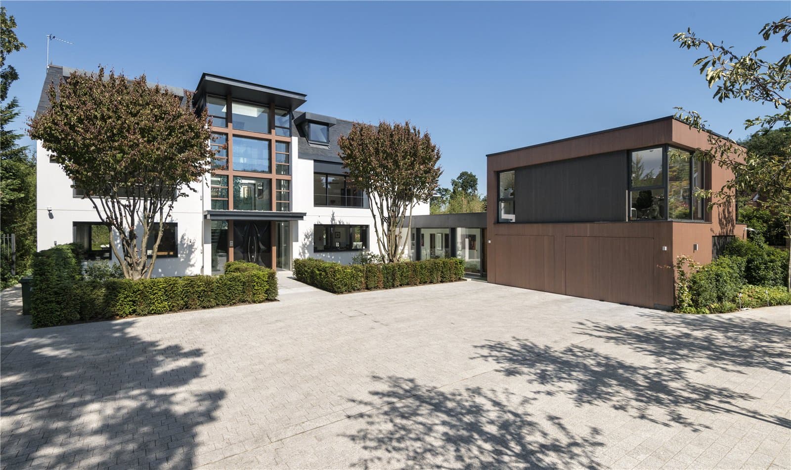 A daytime view of the front elevation, showing a two storey white rendered house with a full height glazed entrance section and a separate dark timber clad garage building. A block paved driveway runs across the front of the property. The Warren, Radlett, Hertfordshire, listed with Prime Property.