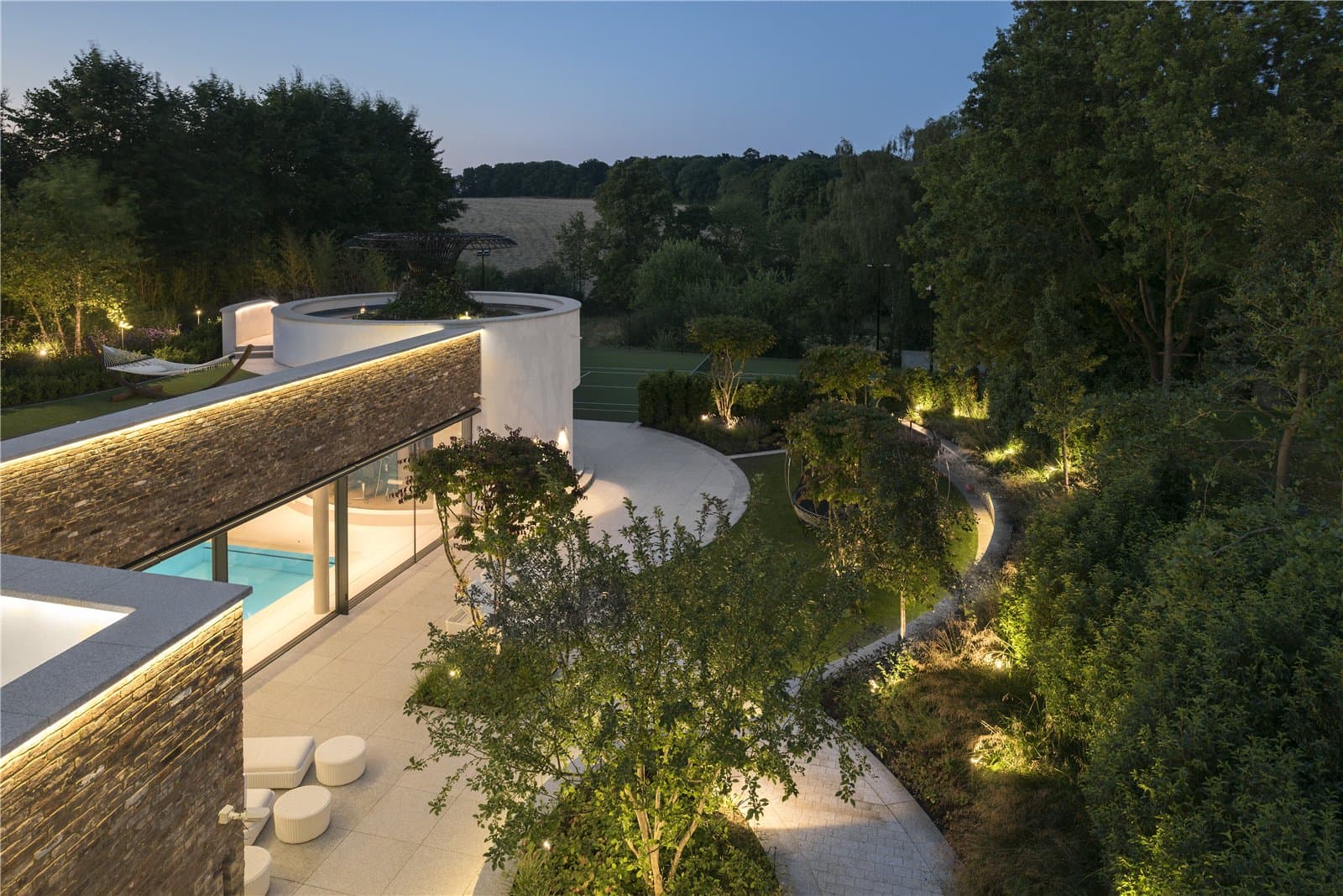 An aerial dusk view of a curved stone terrace and pool building, with landscaped gardens, a circular raised planter and open countryside visible in the background. The Warren, Radlett, Hertfordshire, listed with Prime Property.