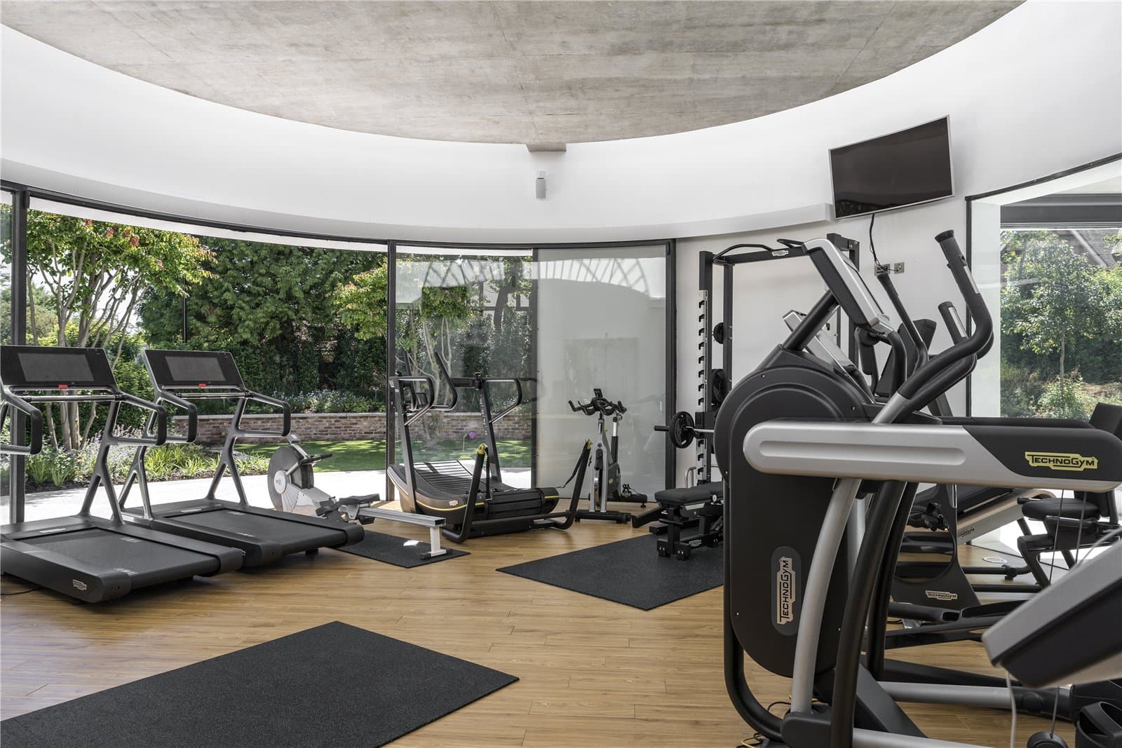 A curved gym space with wood effect flooring, fitted with treadmills, a rowing machine, cross trainer and a multi station weights rack, with floor to ceiling curved glazing overlooking the garden. The Warren, Radlett, Hertfordshire, listed with Prime Property.