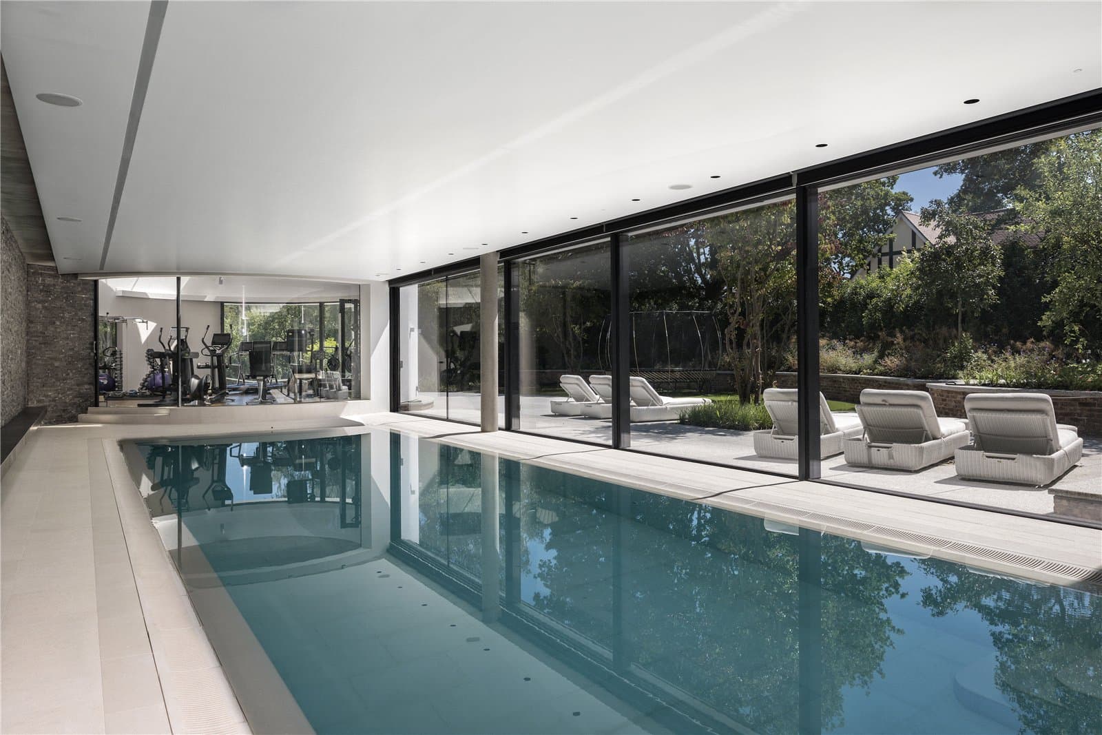 An indoor swimming pool runs alongside full height glass doors leading out to the terrace, with a gym visible through glazing at one end and loungers set outside on the paving. The Warren, Radlett, Hertfordshire, listed with Prime Property.