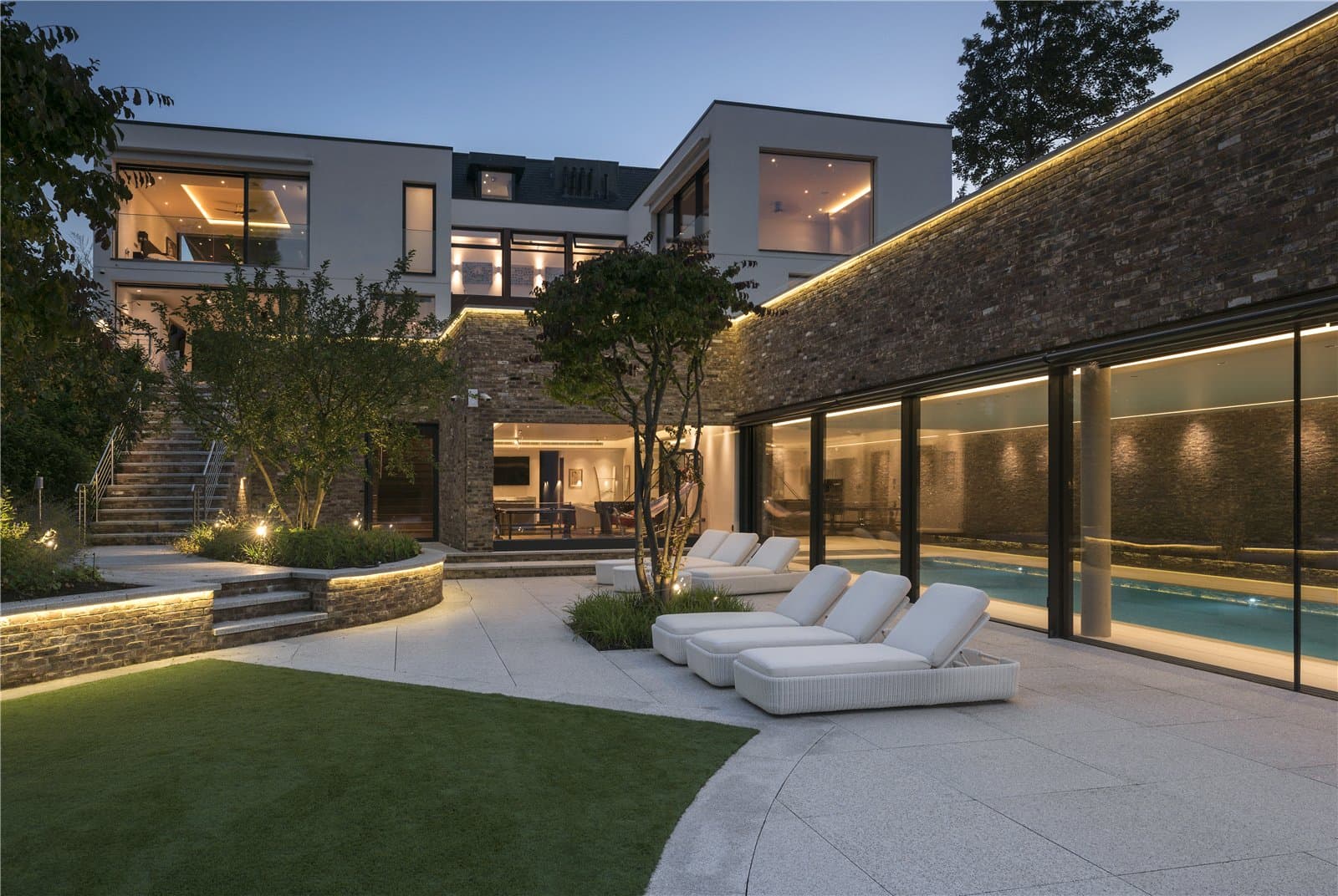 A dusk view of the rear terrace, with a row of white loungers set on a stone patio beside a brick built pool building with full height glass doors. Steps and raised planting lead up to a lawned garden area. The Warren, Radlett, Hertfordshire, listed with Prime Property.