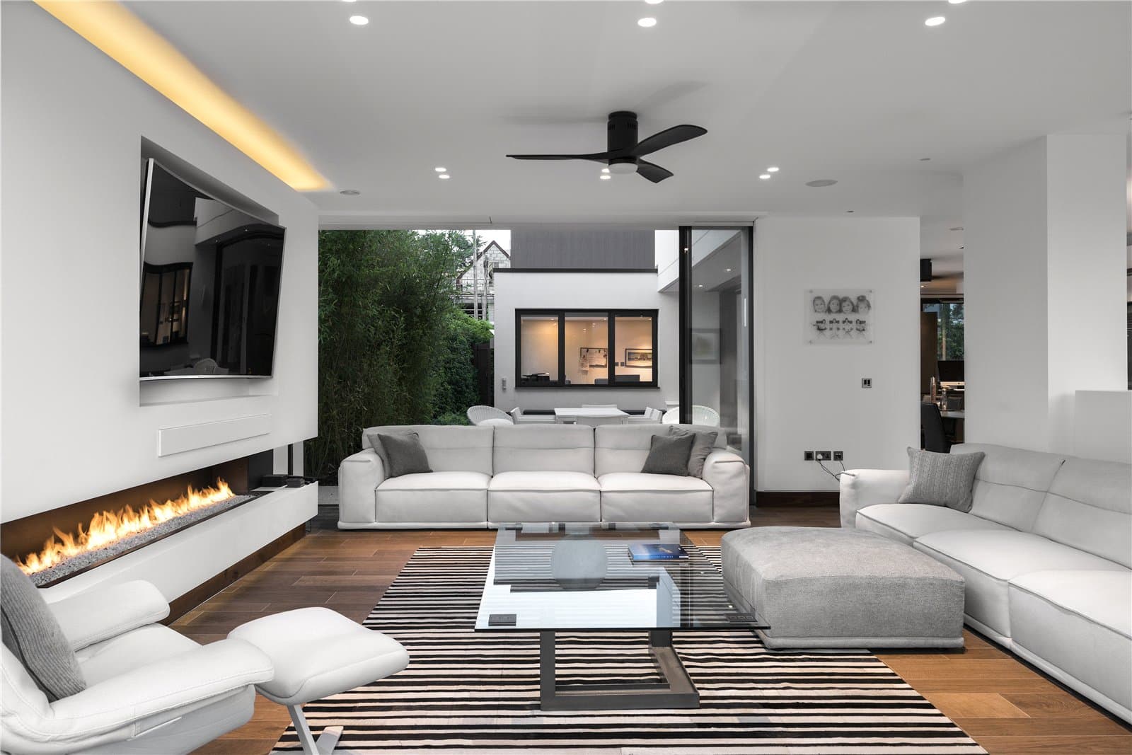A reception room with a built in linear fireplace beneath a wall mounted television, white upholstered sofas and armchair, a glass coffee table and a striped rug over dark wood flooring. The Warren, Radlett, Hertfordshire, listed with Prime Property.