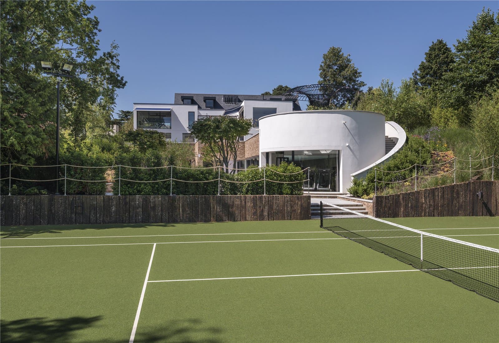 A tennis court bordered by timber sleeper edging and rope fencing, with the main house and a curved white pool building visible beyond, surrounded by mature trees and planting. The Warren, Radlett, Hertfordshire, listed with Prime Property.