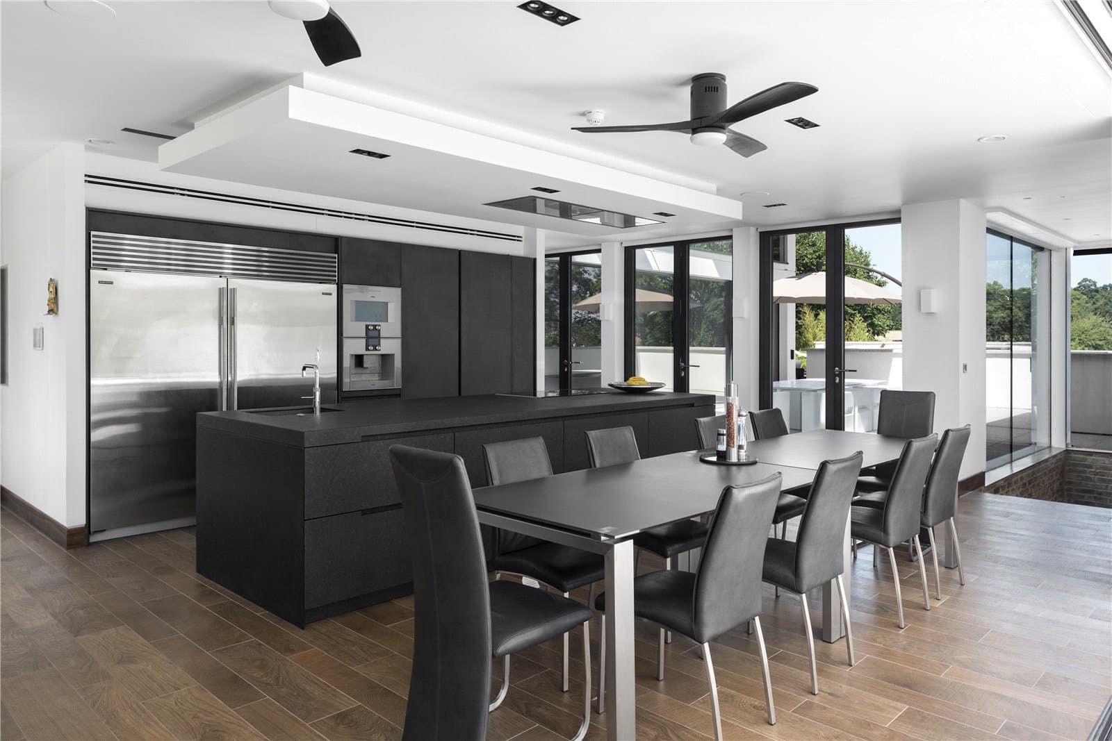A dark toned kitchen with a central island, integrated fridge freezer and built in oven, positioned alongside a long dining table with black leather chairs, with full height windows and doors along one side. The Warren, Radlett, Hertfordshire, listed with Prime Property.